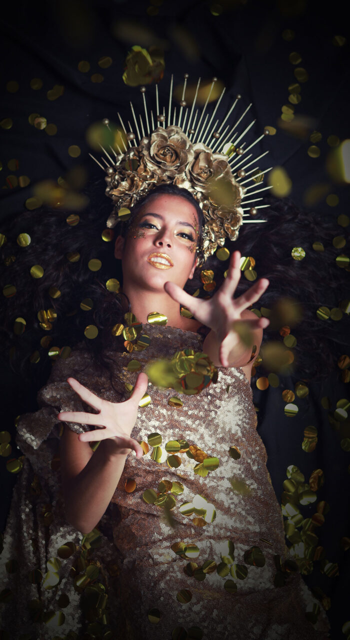 Mujer acostada con corona y vestido, papel dorado cayendo sobre ella.